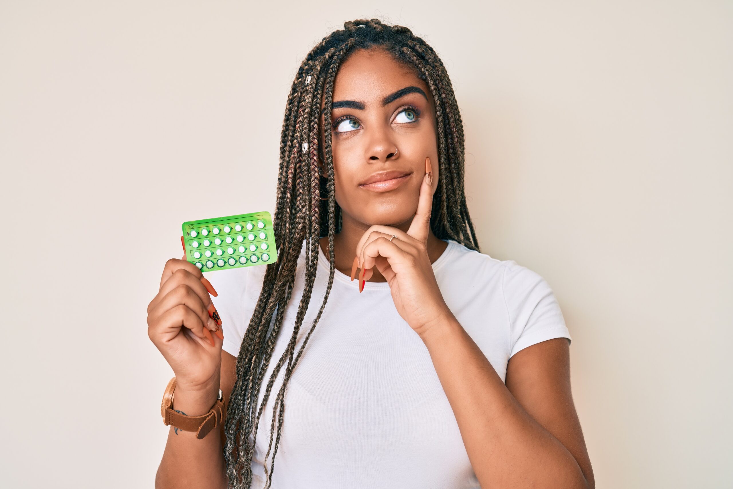 young woman with long braided hair thinking with her finger to her cheek while holding a packet of birth control pills